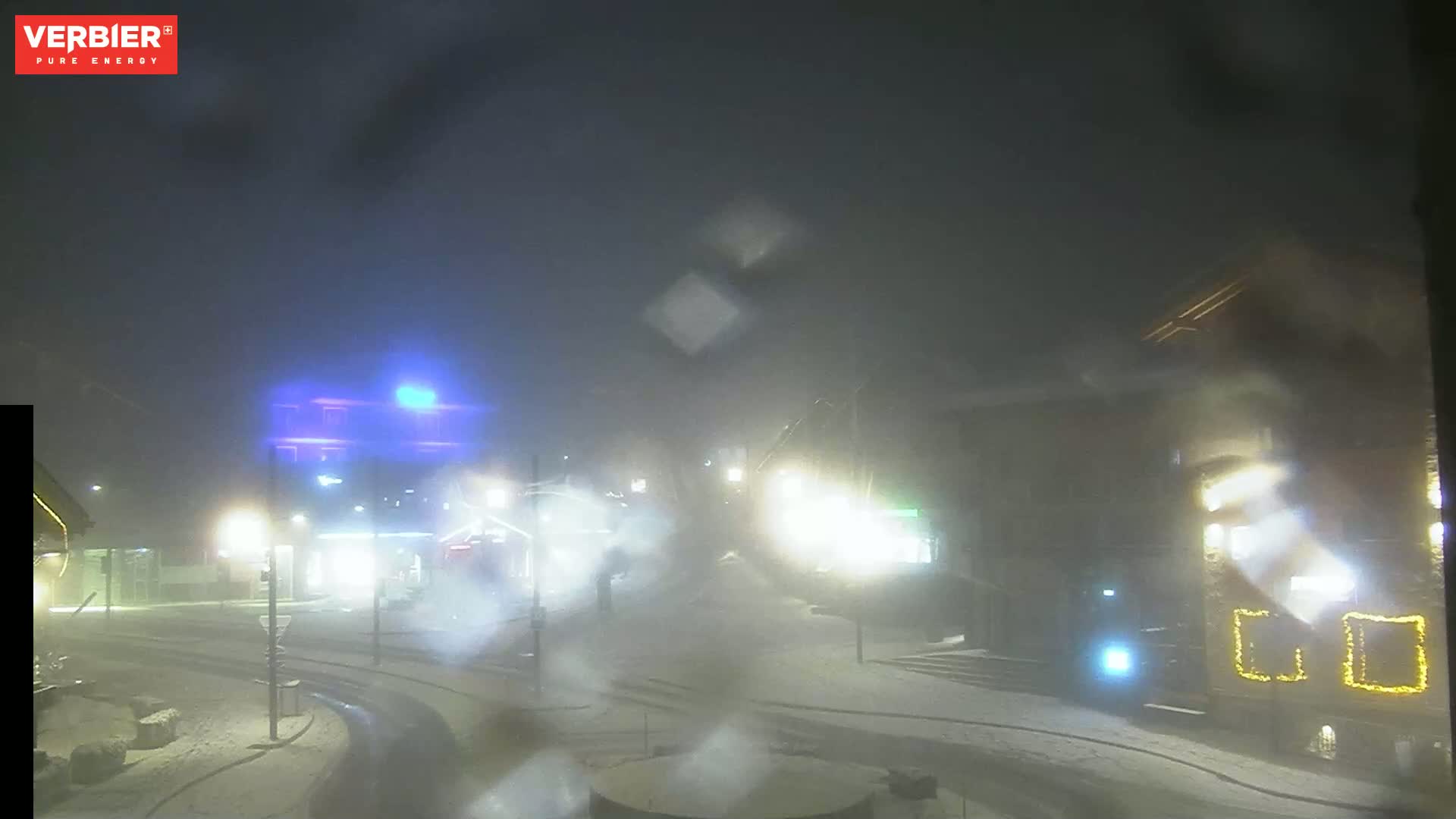 A snow-covered street scene at night, obscured by heavy fog or falling snow, shows dimly lit buildings and vehicles.