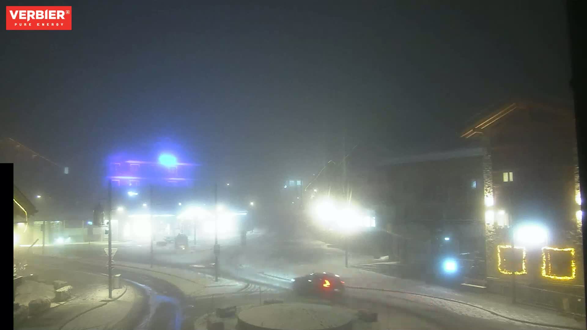 A foggy, snowy night scene shows a street intersection with several buildings and vehicles, illuminated by streetlights.
