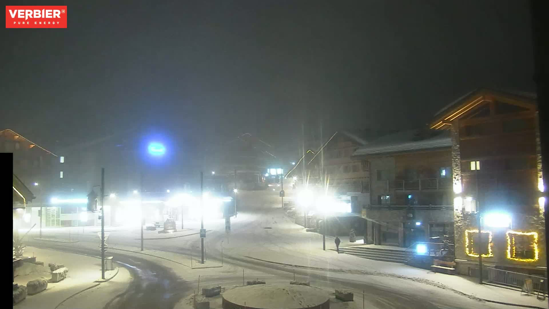 A snow-covered town square at night, illuminated by streetlights in foggy conditions.
