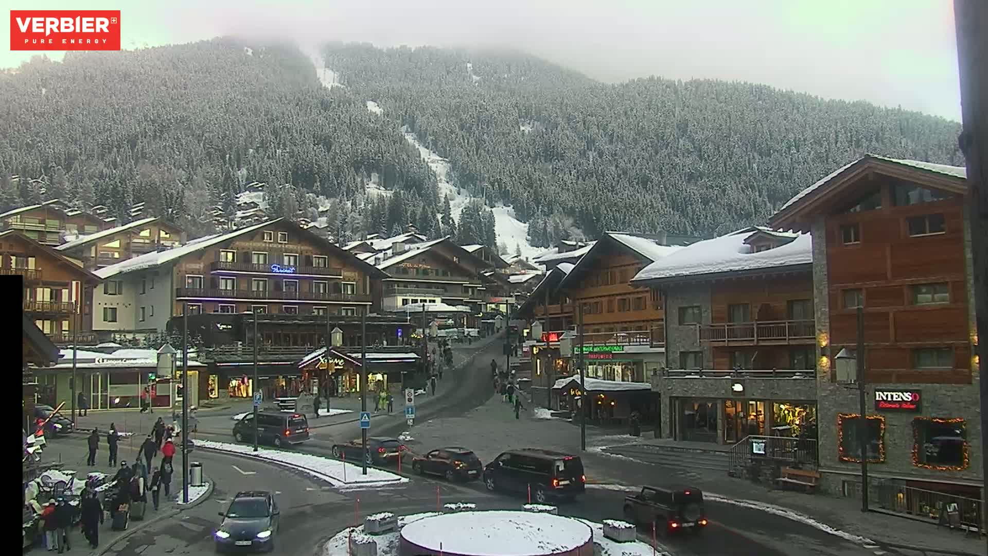 A snow-covered village nestled in a mountain valley on an overcast day, with cars and pedestrians visible in a town square.