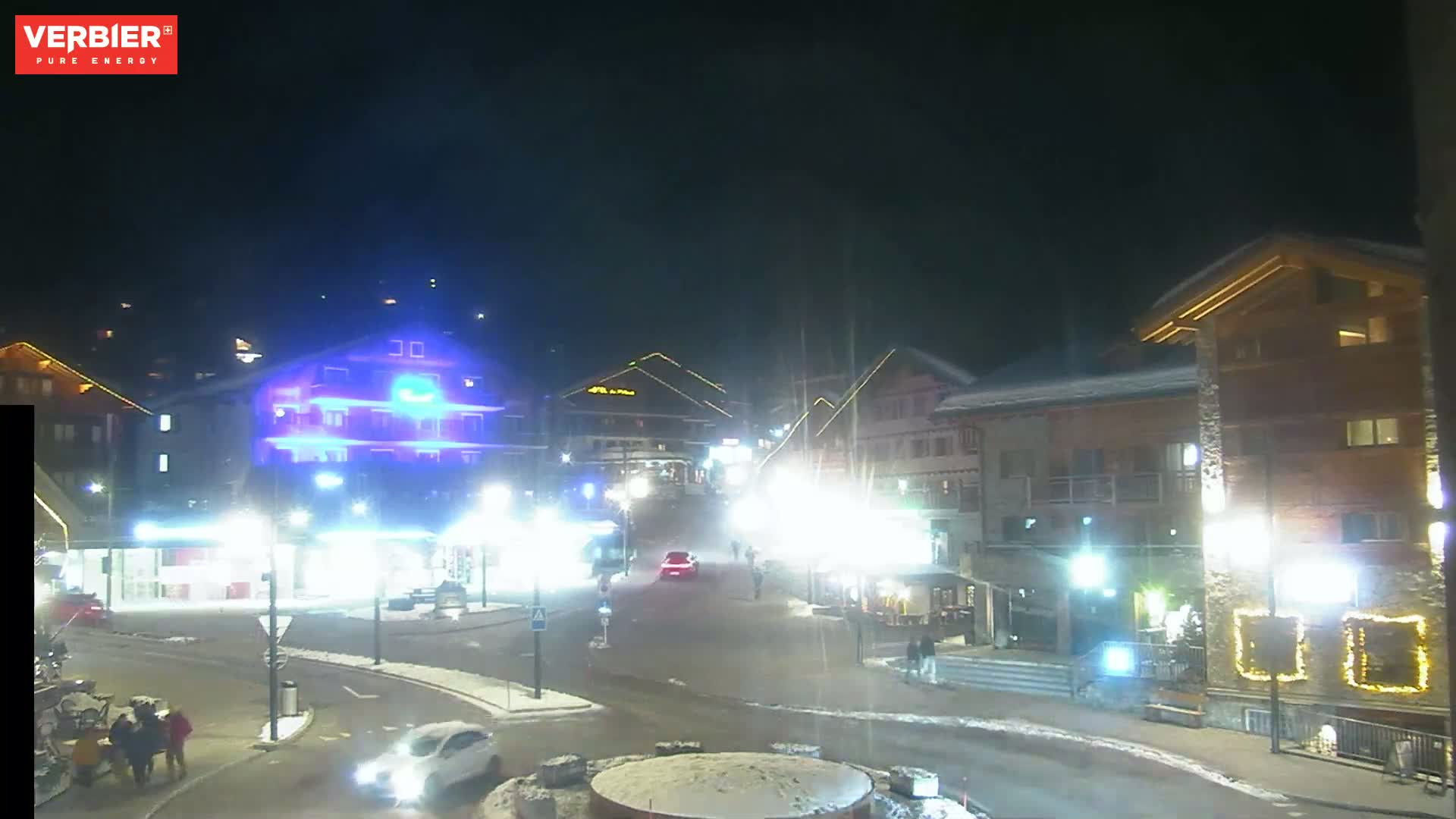 A nighttime scene of a snow-covered town square with brightly lit buildings and a few cars and pedestrians.