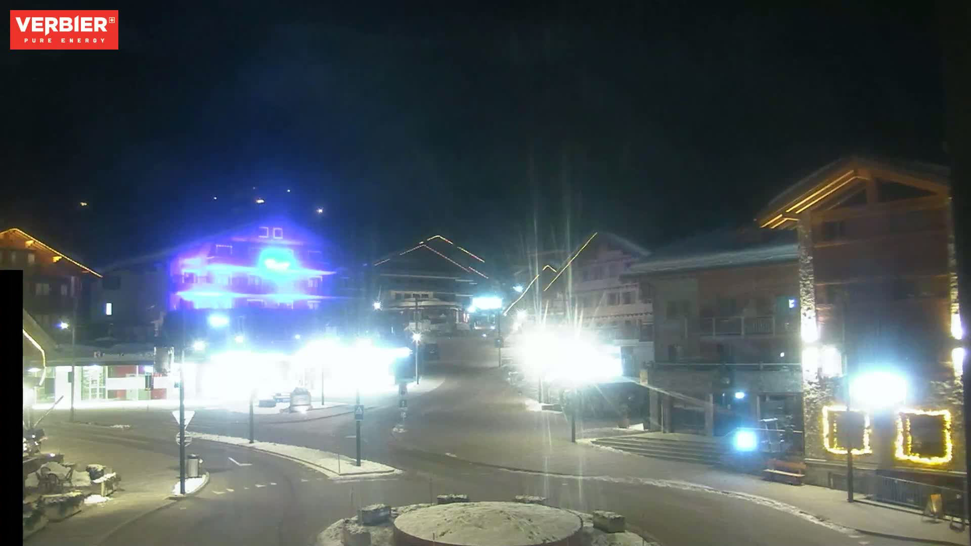 A nighttime view of a snow-covered town square with brightly lit buildings and streetlights under a clear, dark sky.