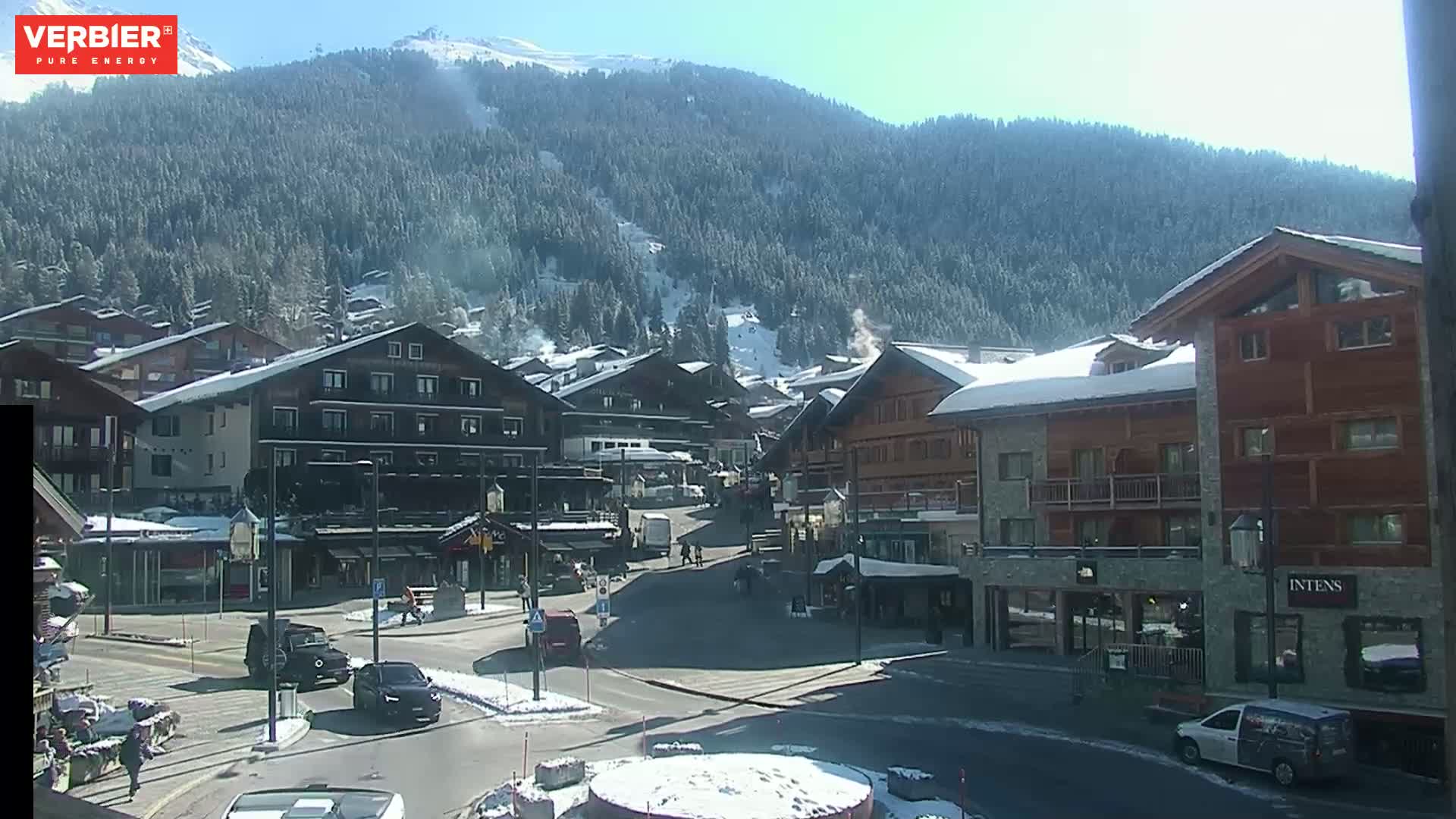 A sunny day in a snow-covered village nestled in a mountain valley, with cars parked along a street in front of several buildings.