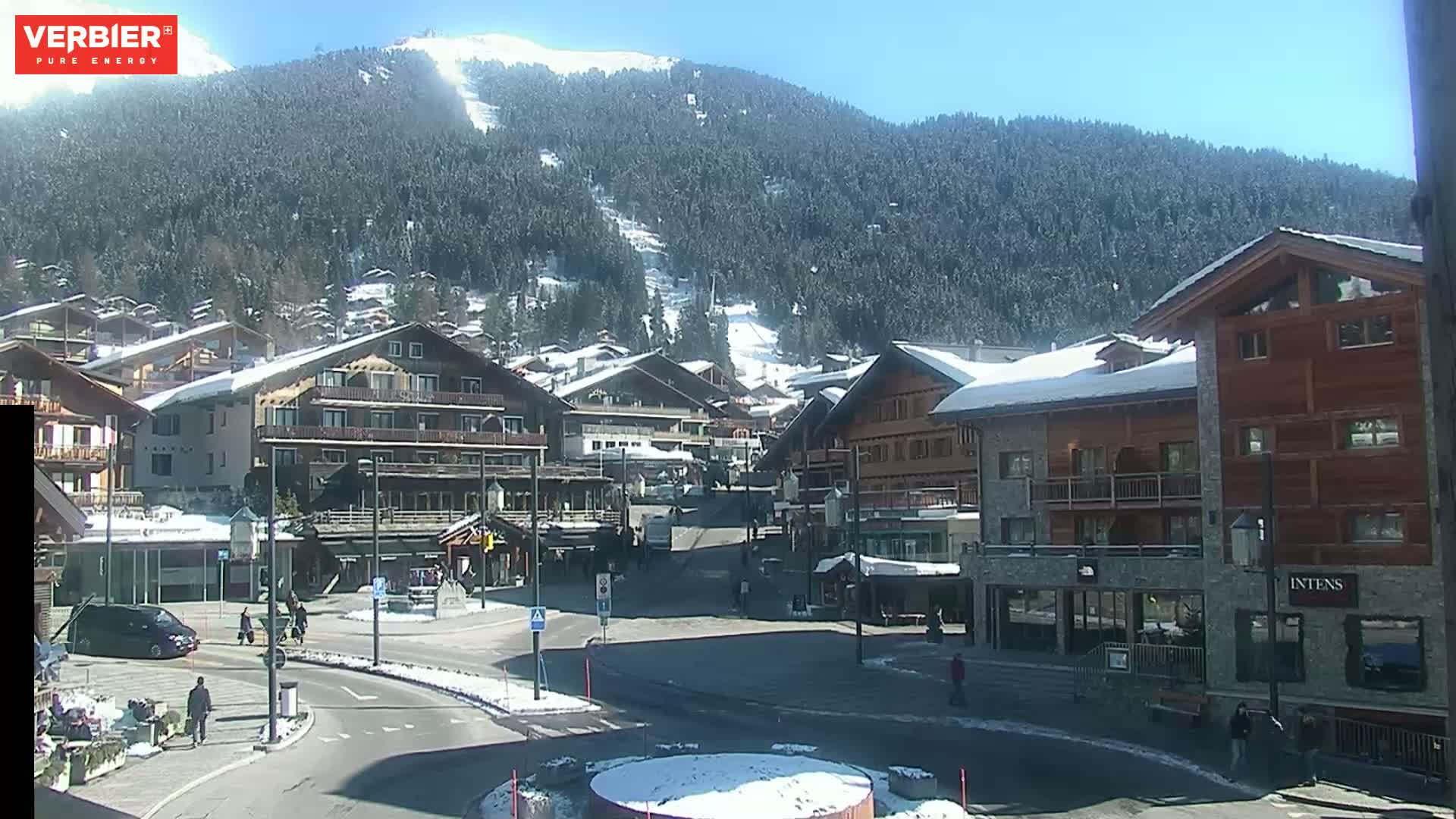 A sunny, snow-covered mountain village square with several multi-story buildings, some with snow on their roofs, and a few pedestrians.