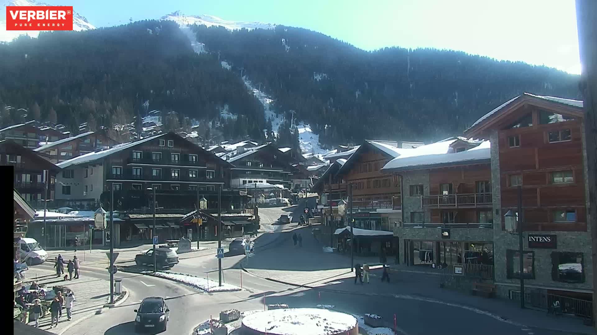 A sunny day in a snow-covered mountain village, showing a town square with several buildings and a few pedestrians.