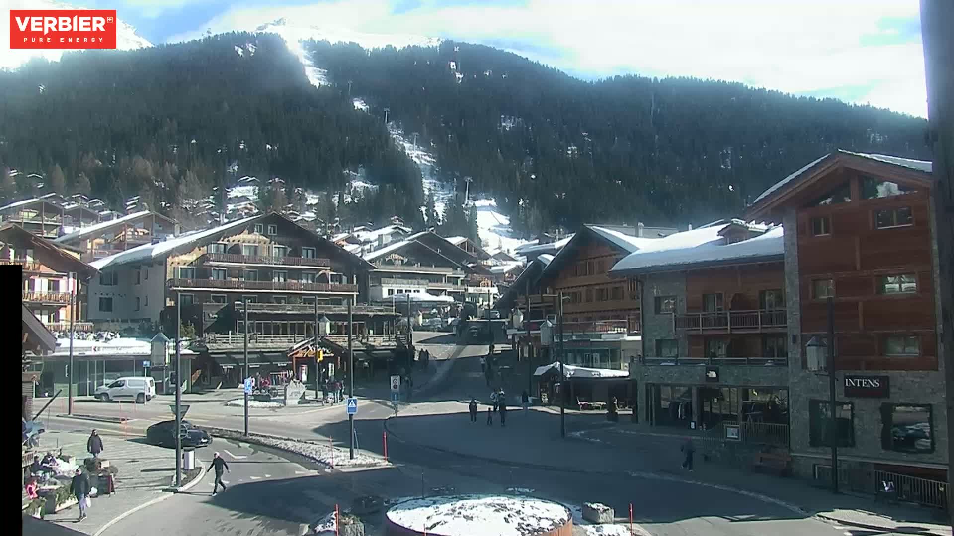 A sunny day in a snow-dusted mountain town shows several buildings surrounding a small plaza with people walking around.