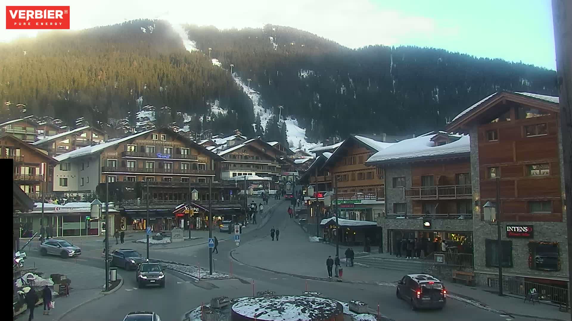 A snow-dusted village nestled in a valley, surrounded by a mountainside with a ski slope, under a mostly clear sky.