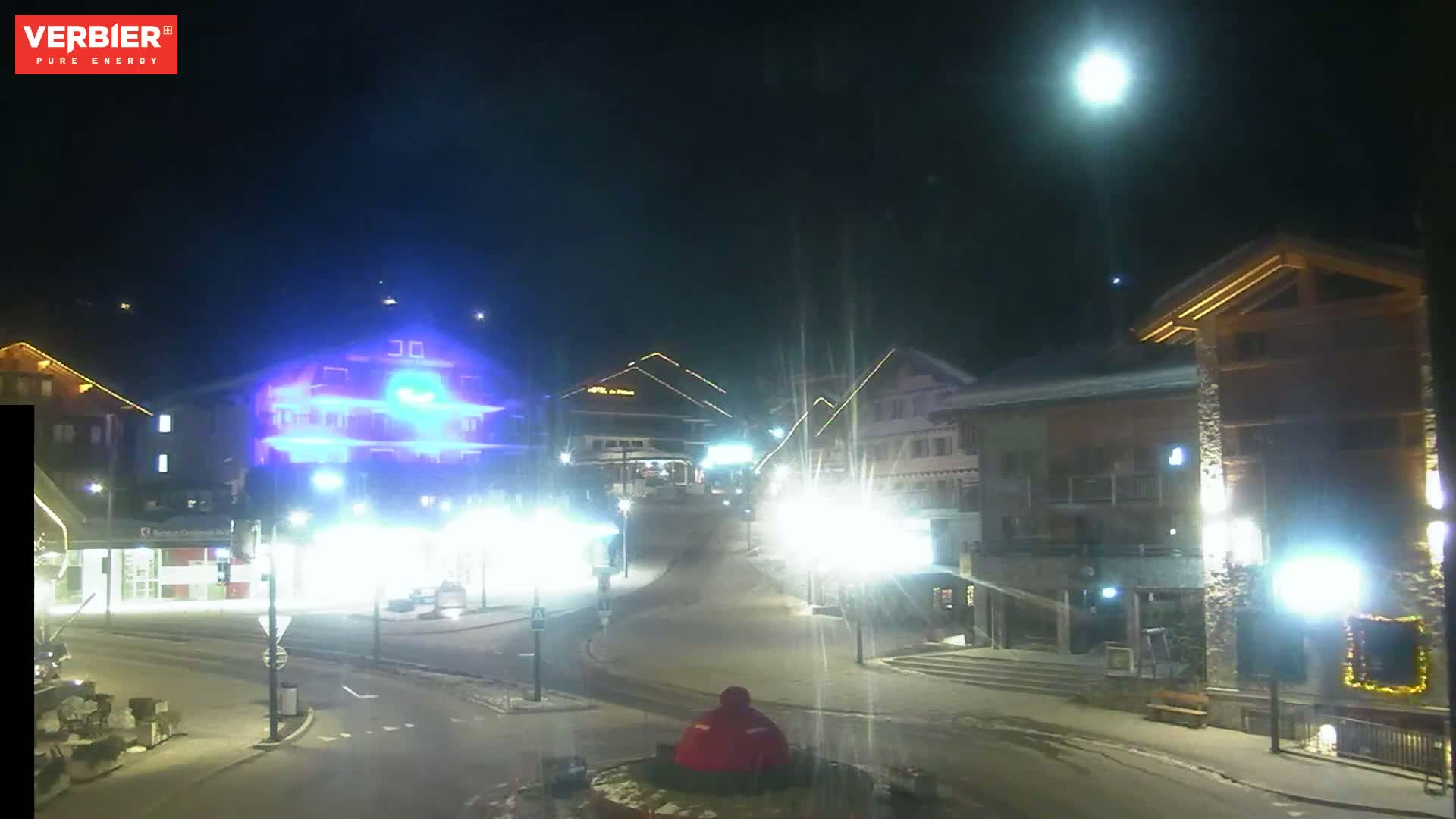 A nighttime view of a snow-covered town square with illuminated buildings under a clear sky and a bright moon.