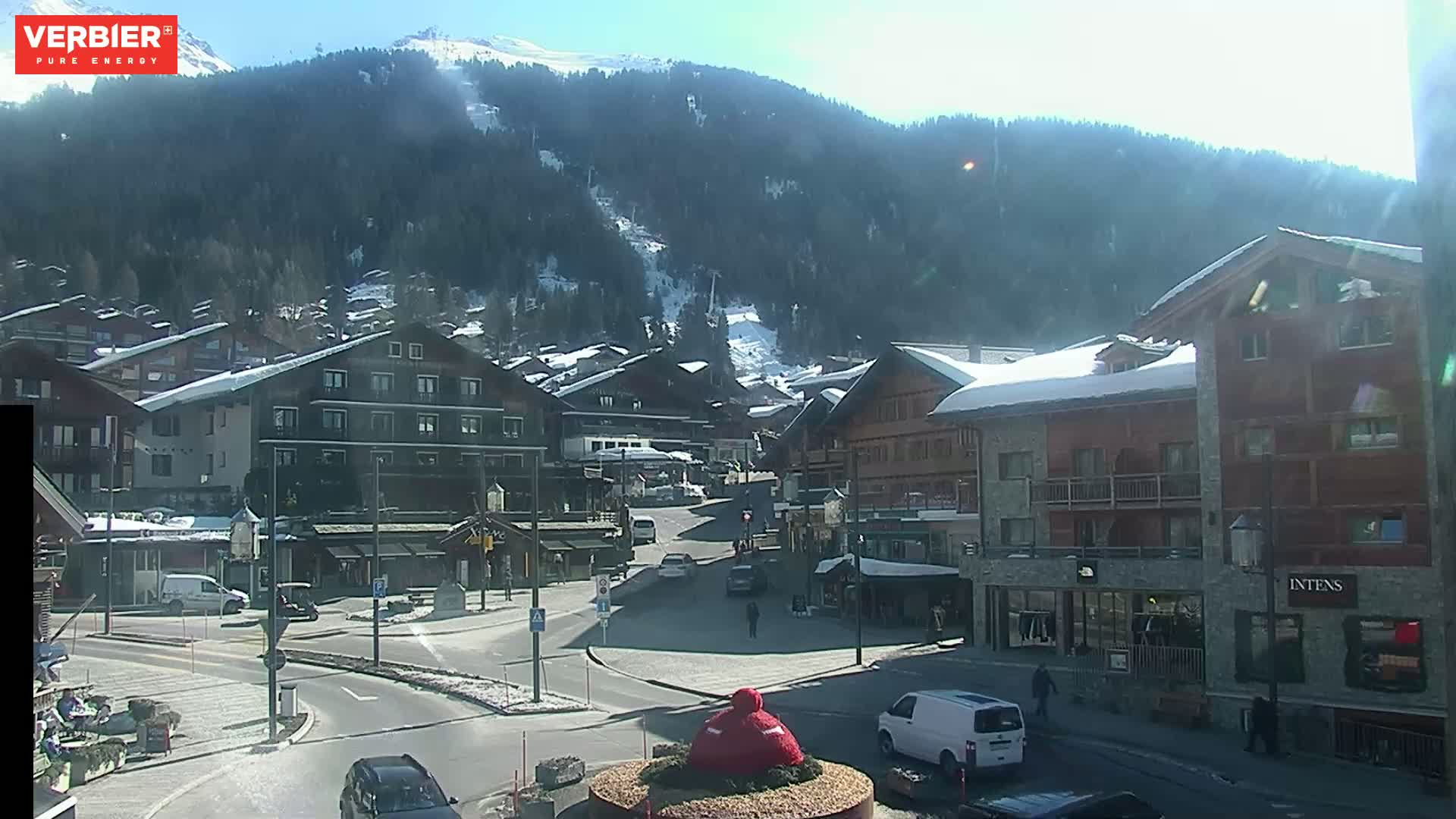A sunny day in a snow-dusted mountain town shows a town square with several buildings, cars, and pedestrians, with a snow-capped mountain in the background.