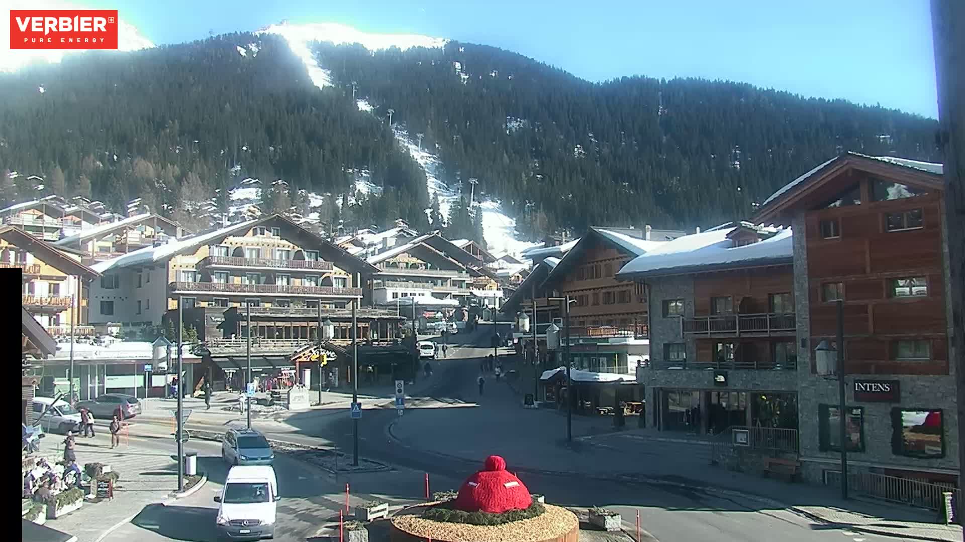 A sunny day reveals a village nestled in a snow-covered mountain valley, with several buildings and cars visible in a town square.