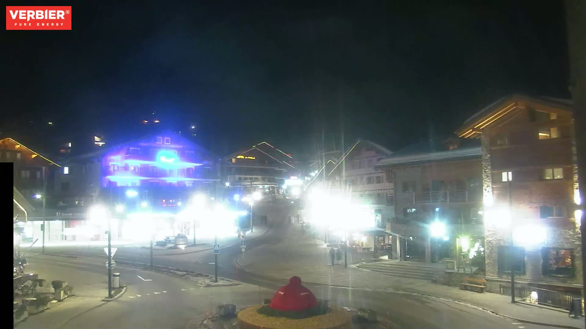 A nighttime view of a town square with several illuminated buildings, streetlights, and a small, red object in the center of a roundabout.