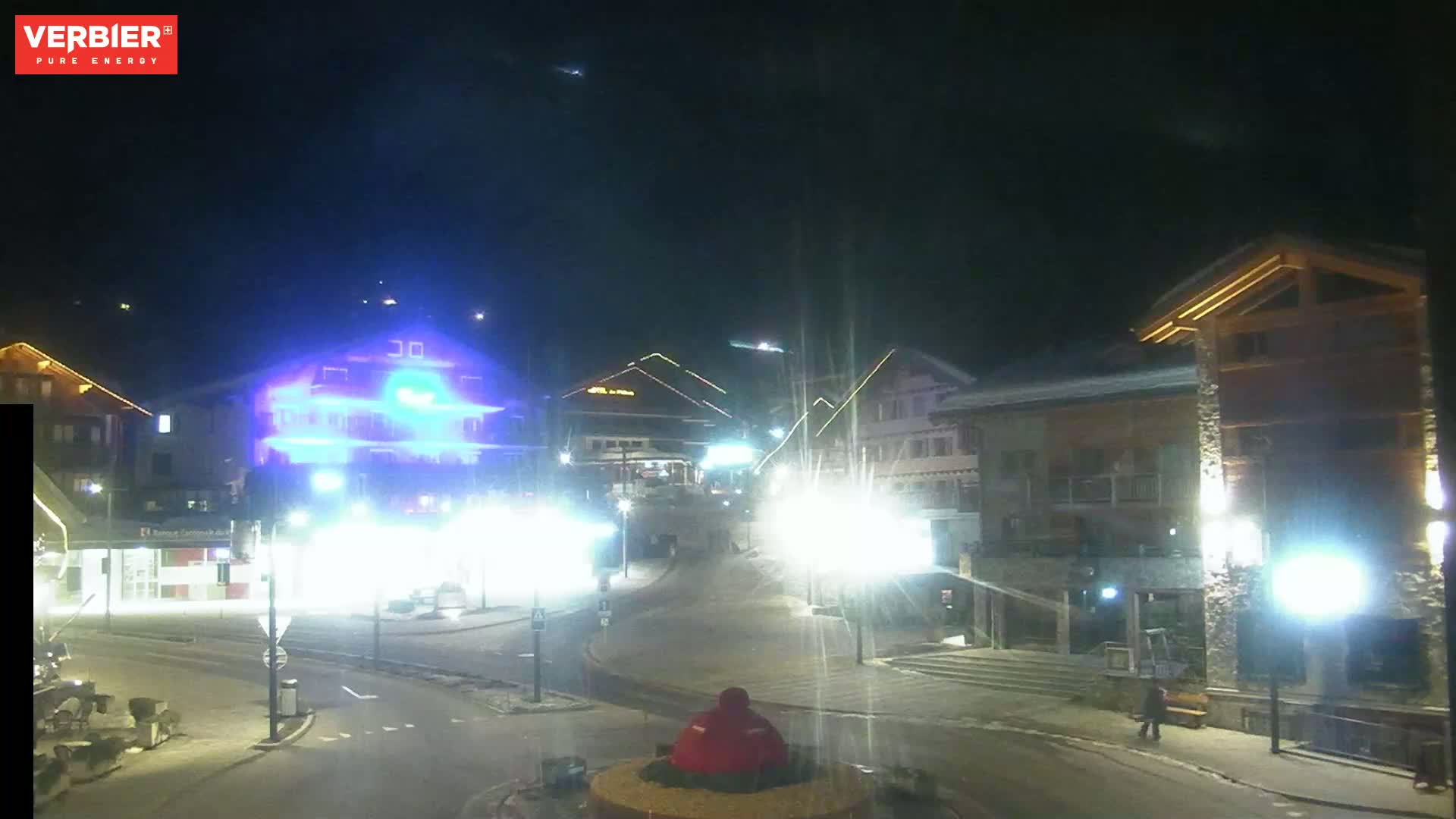 A nighttime view of a snow-covered town square with brightly lit buildings and streetlights.