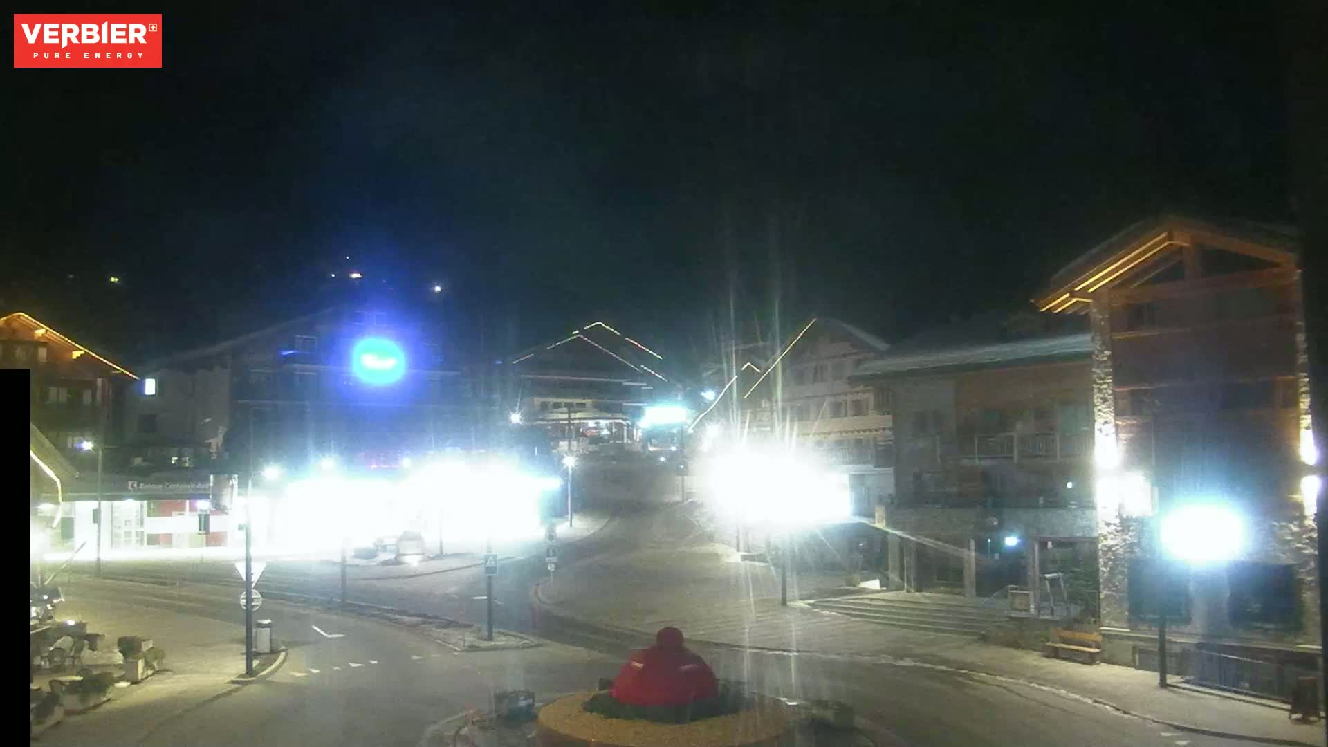 A nighttime view of a snow-covered town square with illuminated buildings and streetlights.