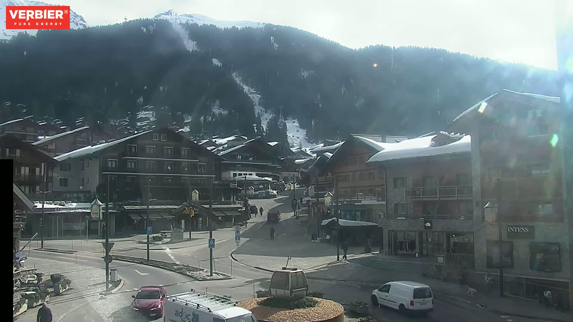 A sunny day in a snow-covered mountain village shows a town square with buildings, vehicles, and a few pedestrians, with a mountain backdrop partially covered in snow.