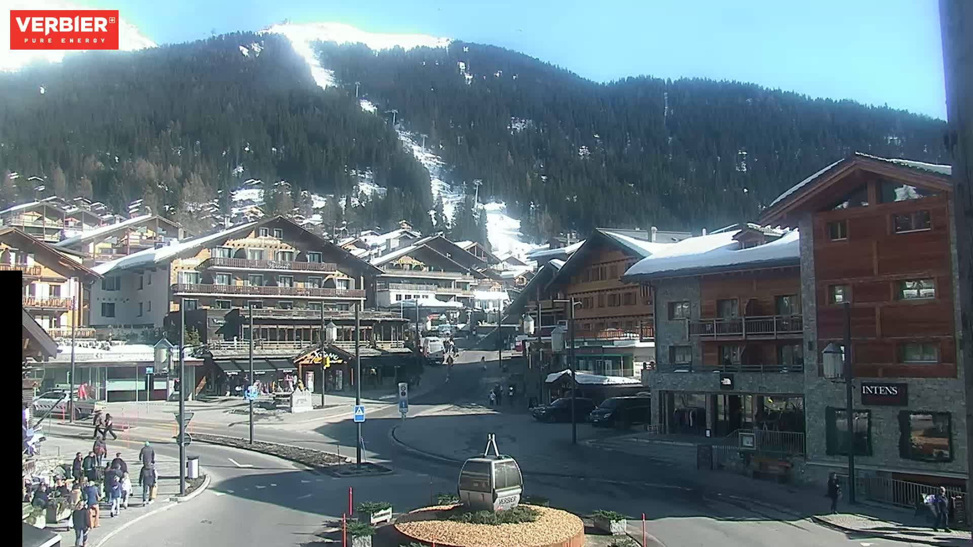 A sunny day in a snow-covered mountain village, showing several multi-story buildings, a roadway, pedestrians, and a ski slope visible in the background.