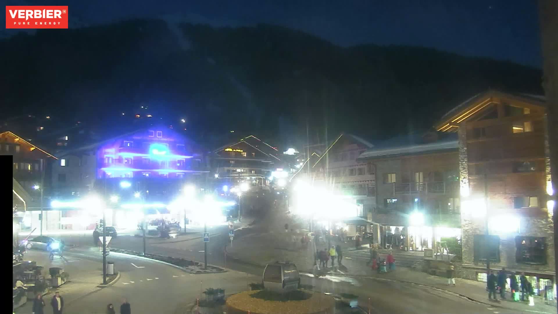 A nighttime view of a snow-covered town square with illuminated buildings and people walking around, under a dark sky.