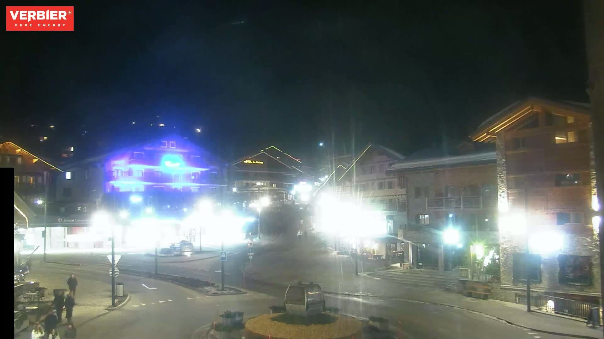 A nighttime view of a snow-covered town square with brightly lit buildings and a few pedestrians.