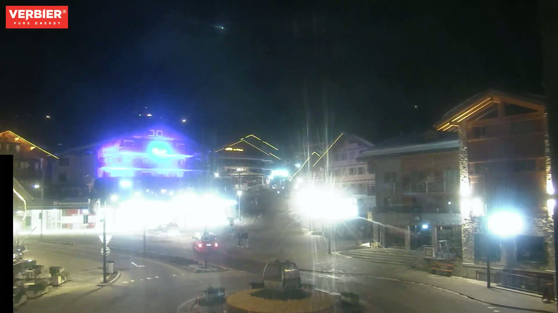 A nighttime, snow-covered town square is illuminated by streetlights, showcasing several buildings and a few vehicles.