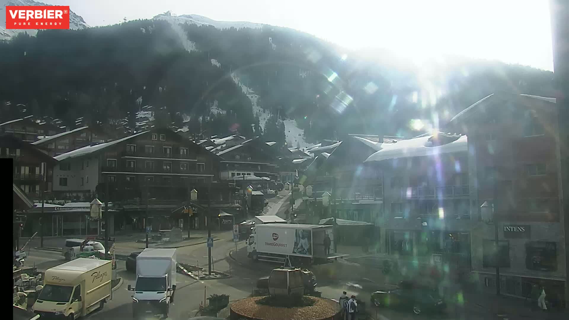 A sunny, snow-covered mountain village scene with several vehicles on a street, viewed from a slightly elevated perspective.