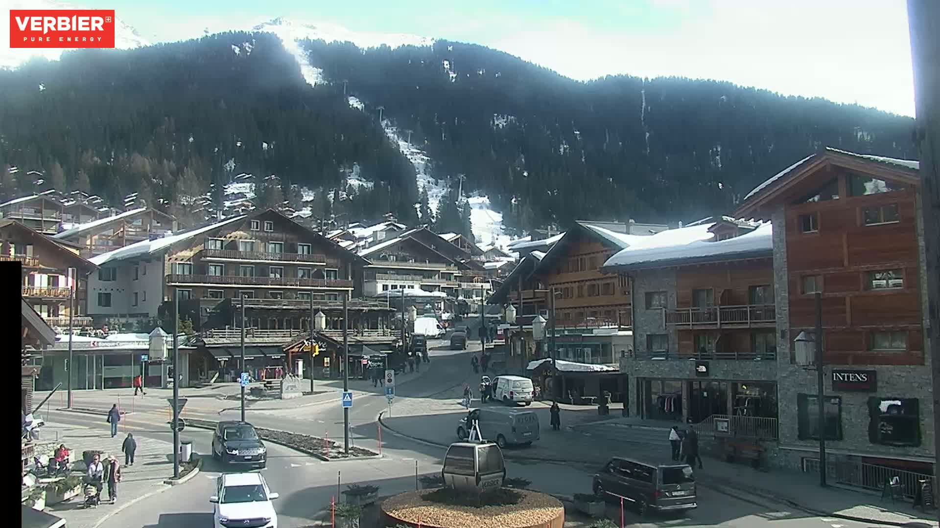 A sunny day in a snow-covered mountain village shows a town square with cars, pedestrians, and buildings, with a ski slope visible on a nearby mountain.