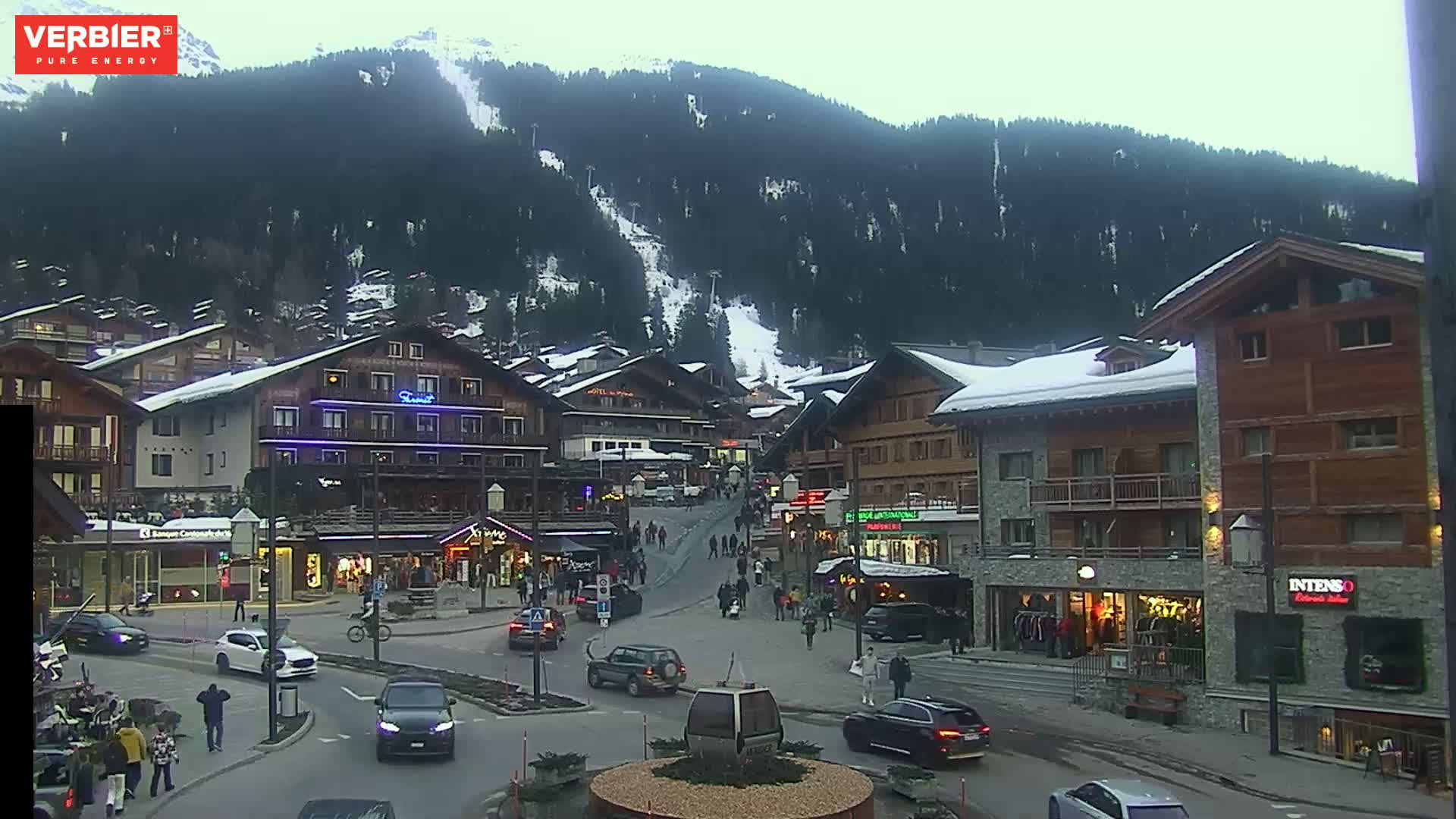A snow-dusted village nestled in a valley surrounded by a dark, snow-capped mountain range under an overcast sky.