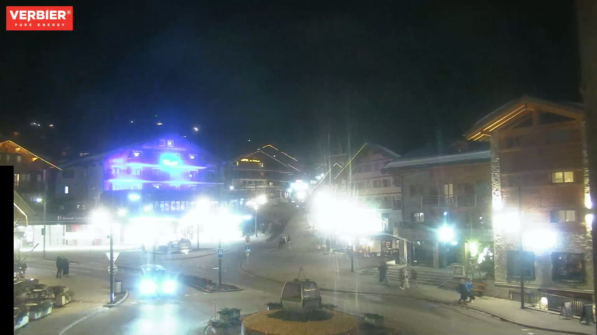 A nighttime, brightly lit town square with several buildings and people walking around under a clear, dark sky.