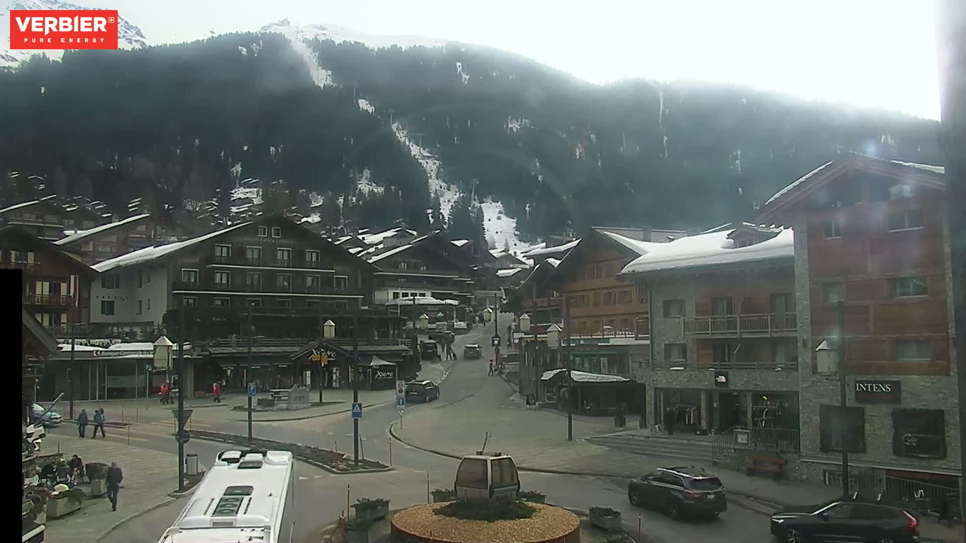 A mountain village scene on an overcast day shows a street with cars and pedestrians, buildings with snow on their roofs, and a mountainside with ski runs in the background.