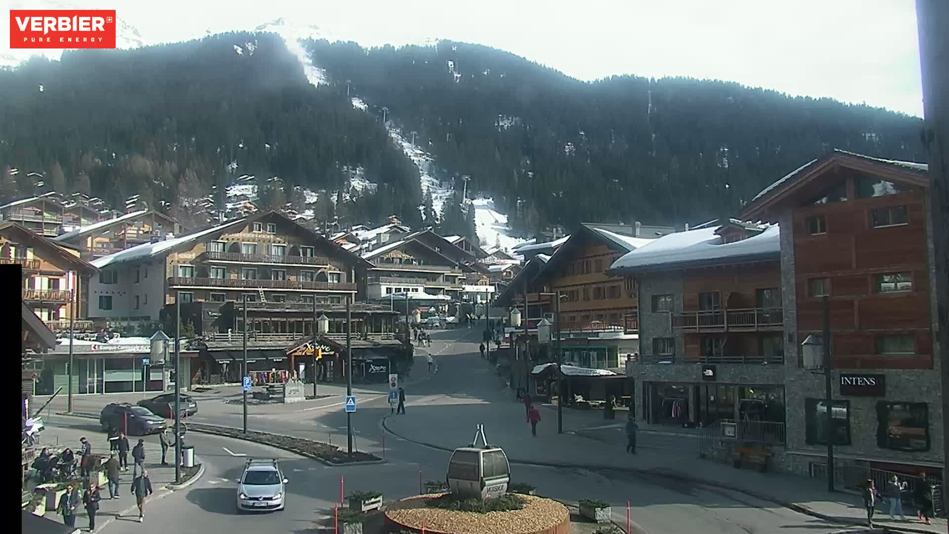 A mostly sunny day in a snow-dusted alpine village with several buildings, a few cars, and pedestrians on the street, a ski slope visible in the background.