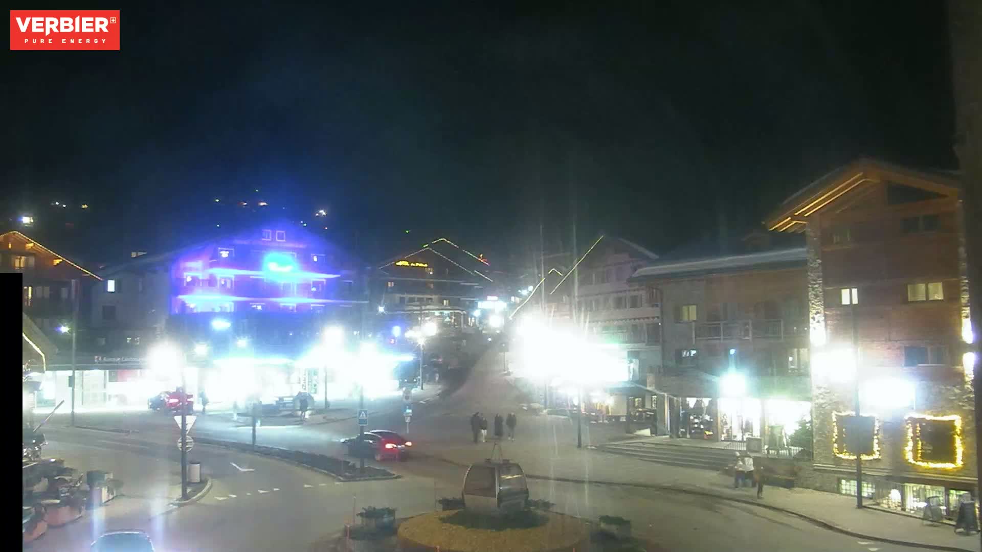 A nighttime view of a town square with several buildings illuminated, a gondola lift in the center, and a few cars and pedestrians.