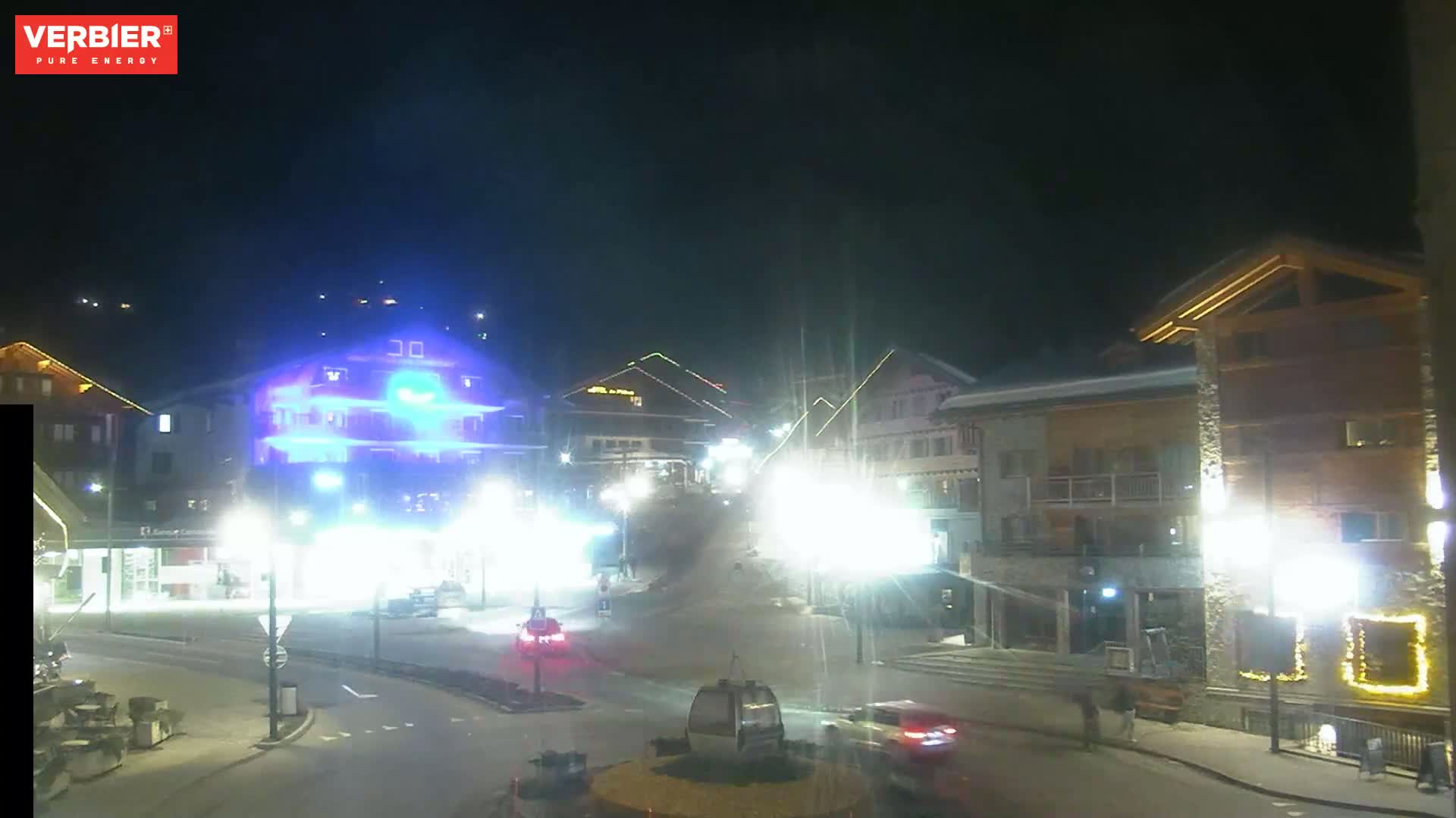 A brightly lit town square at night, with several buildings illuminated, cars on the road, and a cable car in the center.