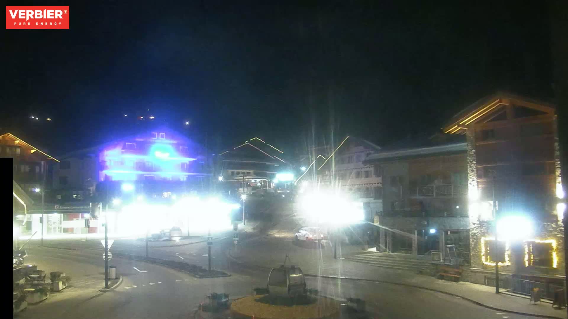 A nighttime view of a town square with brightly lit buildings and a few vehicles, under a clear, dark sky.