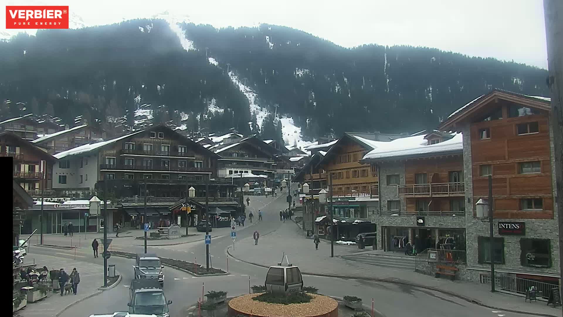 A mostly empty town square in a snow-dusted mountain village on an overcast day, with a ski slope visible on the mountainside in the background.