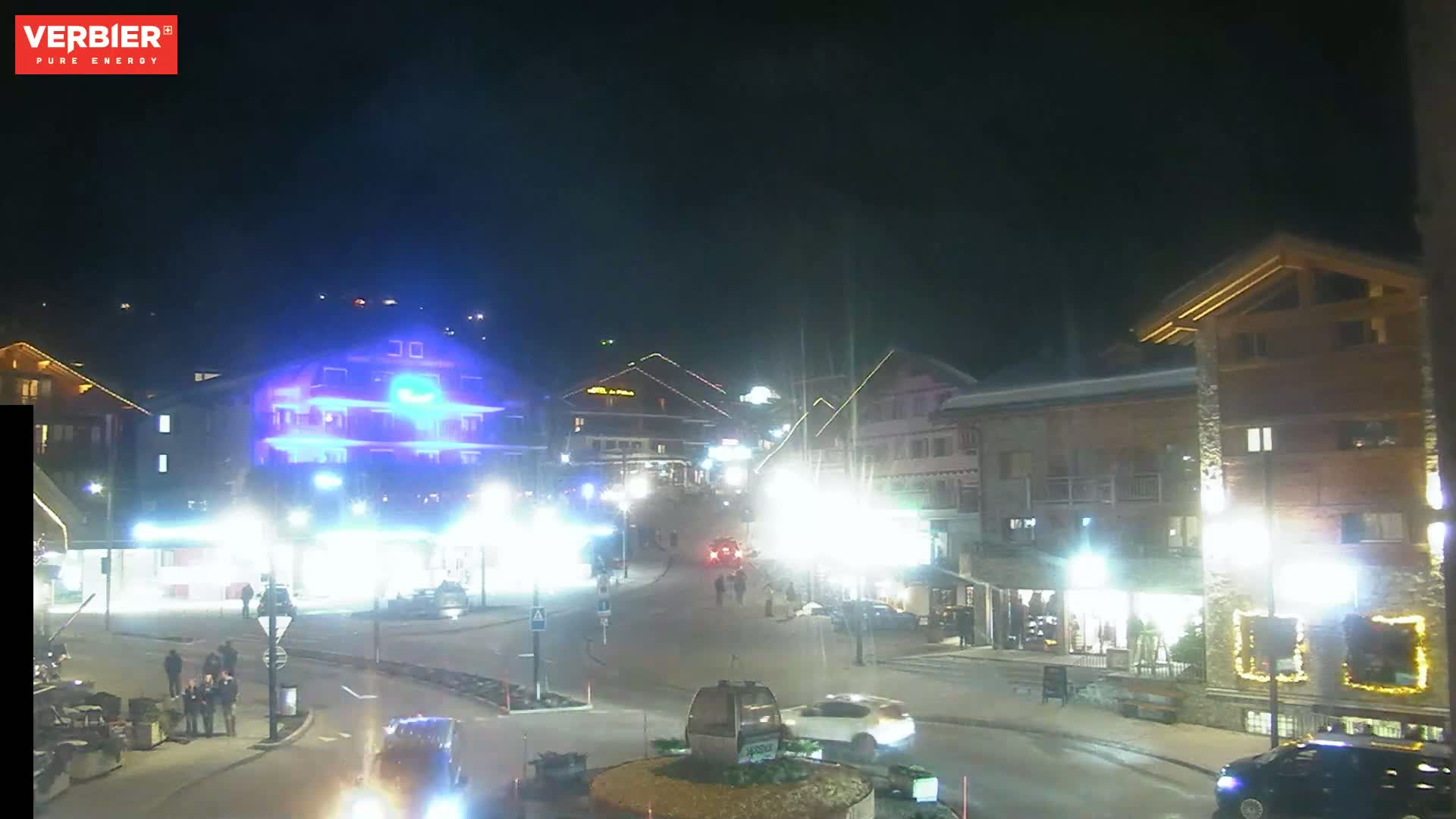 A nighttime street scene shows a town square with buildings illuminated by streetlights and other lights, some cars, and a few pedestrians.