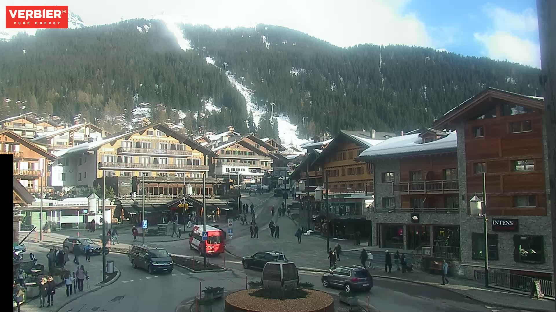 A sunny day in a snow-dusted mountain village with several buildings, pedestrians, and vehicles in a town square, with a ski slope visible on the mountainside.
