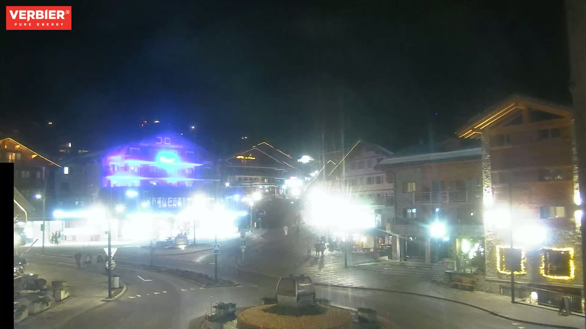 A brightly lit town square at night, with several buildings illuminated, and a cable car gondola in the center.