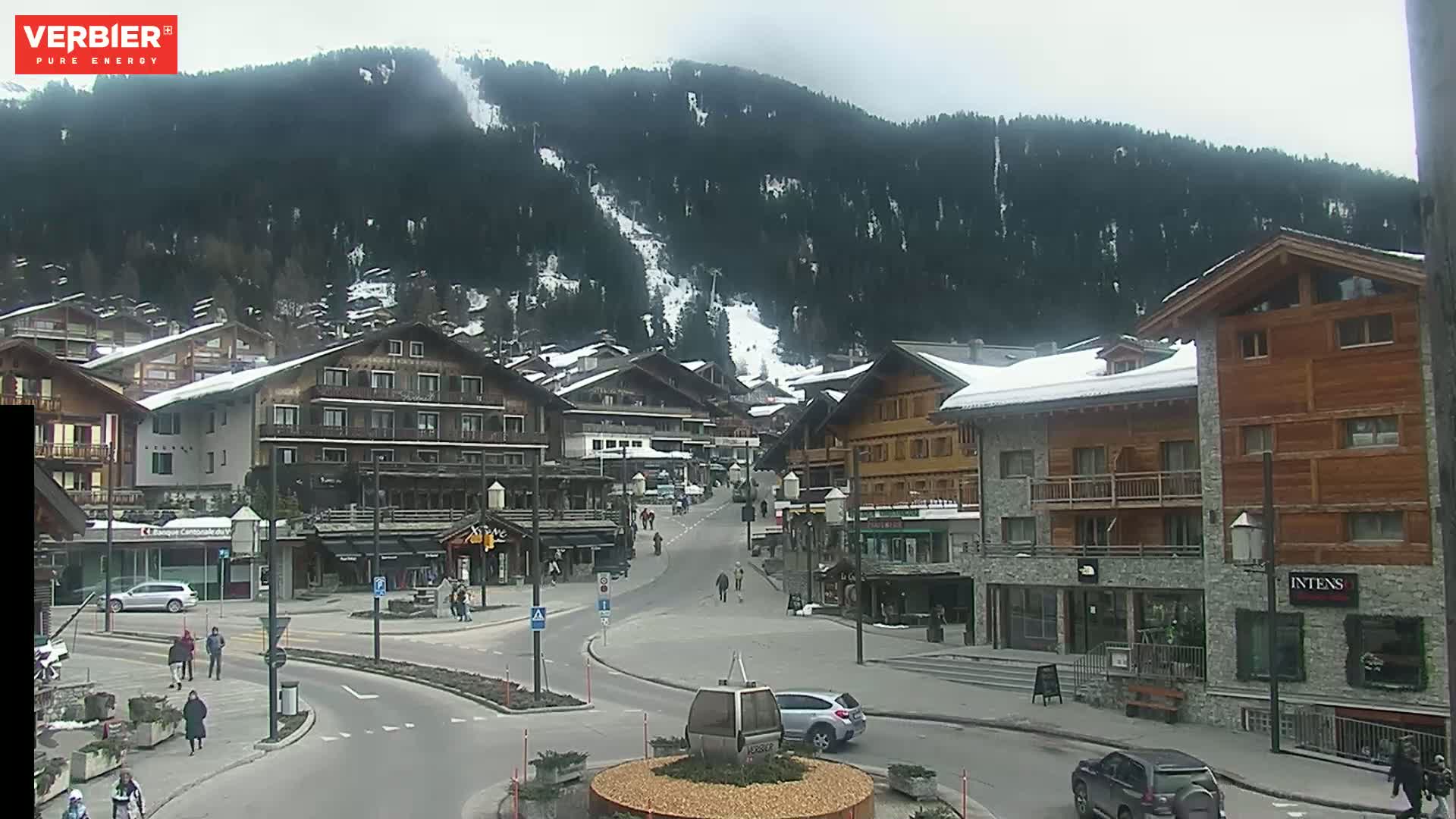 A mostly clear, overcast day reveals a village nestled in a valley, with snow-capped buildings and a ski slope visible on the mountainside.