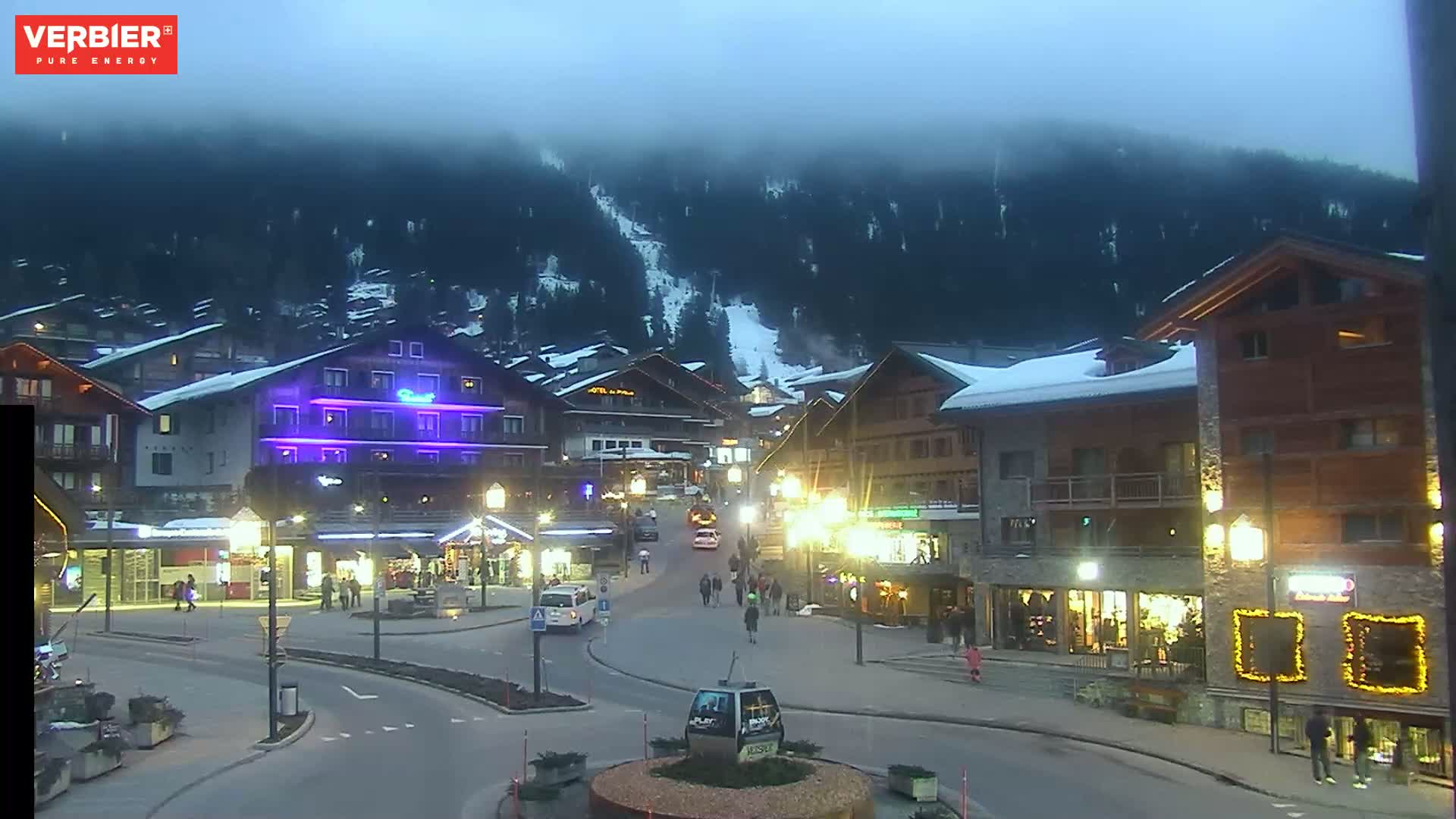 A town square in a snow-covered mountain village is dimly lit at night under a foggy sky.