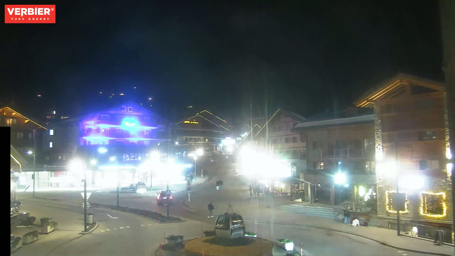 A nighttime view of a town square with brightly lit buildings and a few people walking around, under a dark sky.