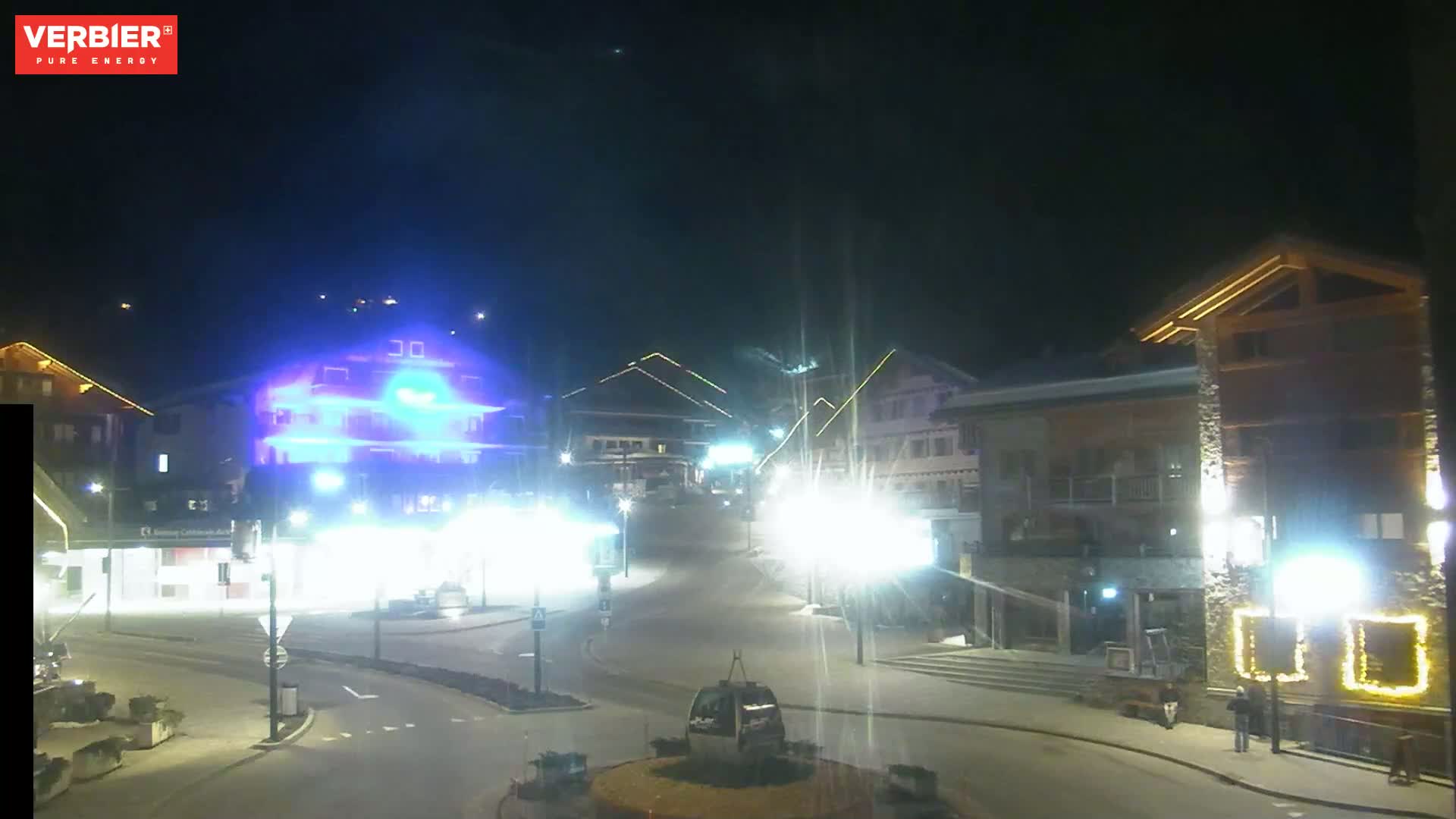 A nighttime view of a town square with brightly lit buildings and a cable car, under a clear, dark sky.