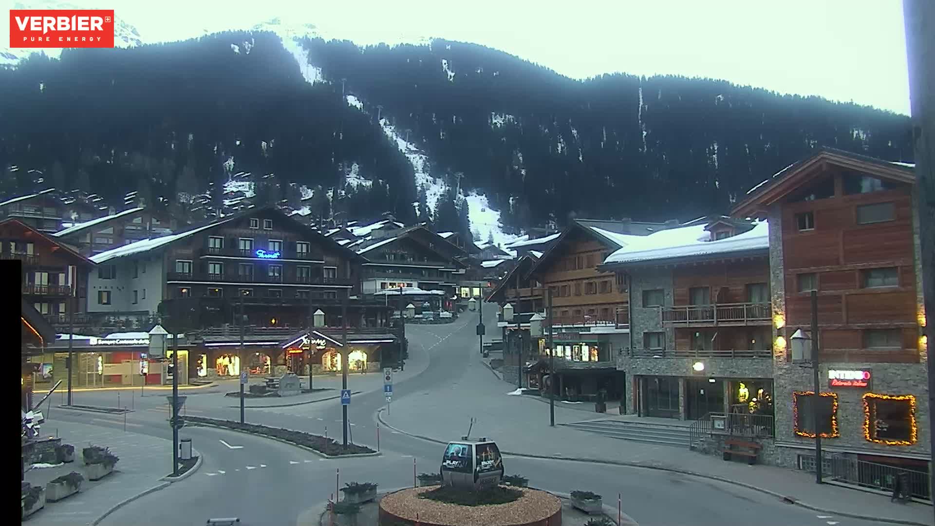 A mostly empty town square in a snow-covered mountain village under an overcast sky.