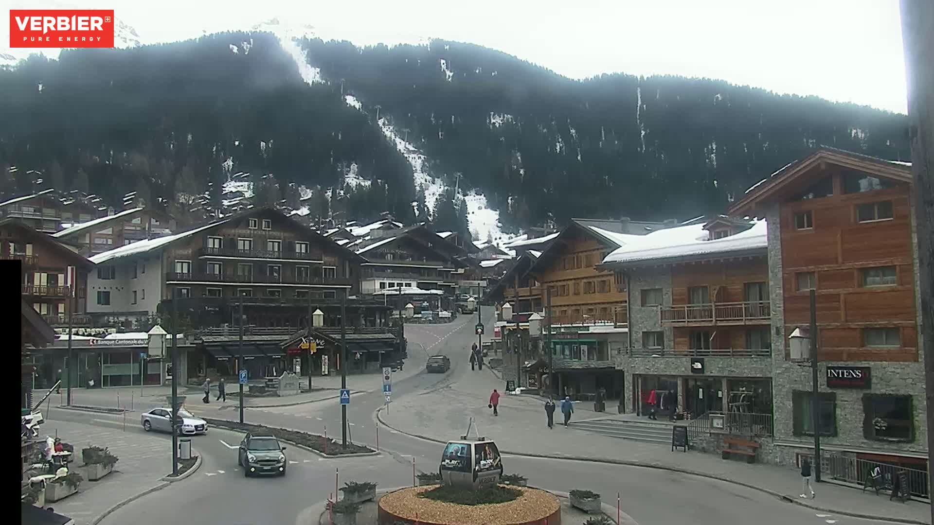 A snow-dusted mountain village street scene on an overcast day, with several buildings and a few cars and pedestrians visible.