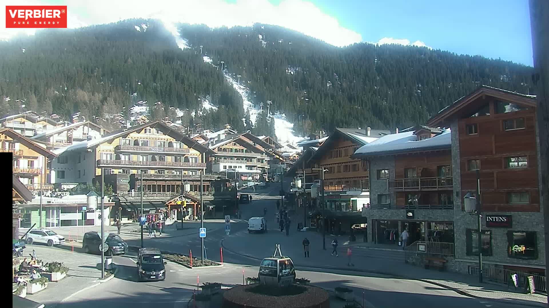 A sunny day in a village nestled in a mountain valley, with several buildings lining a town square and snow-capped mountains in the background.