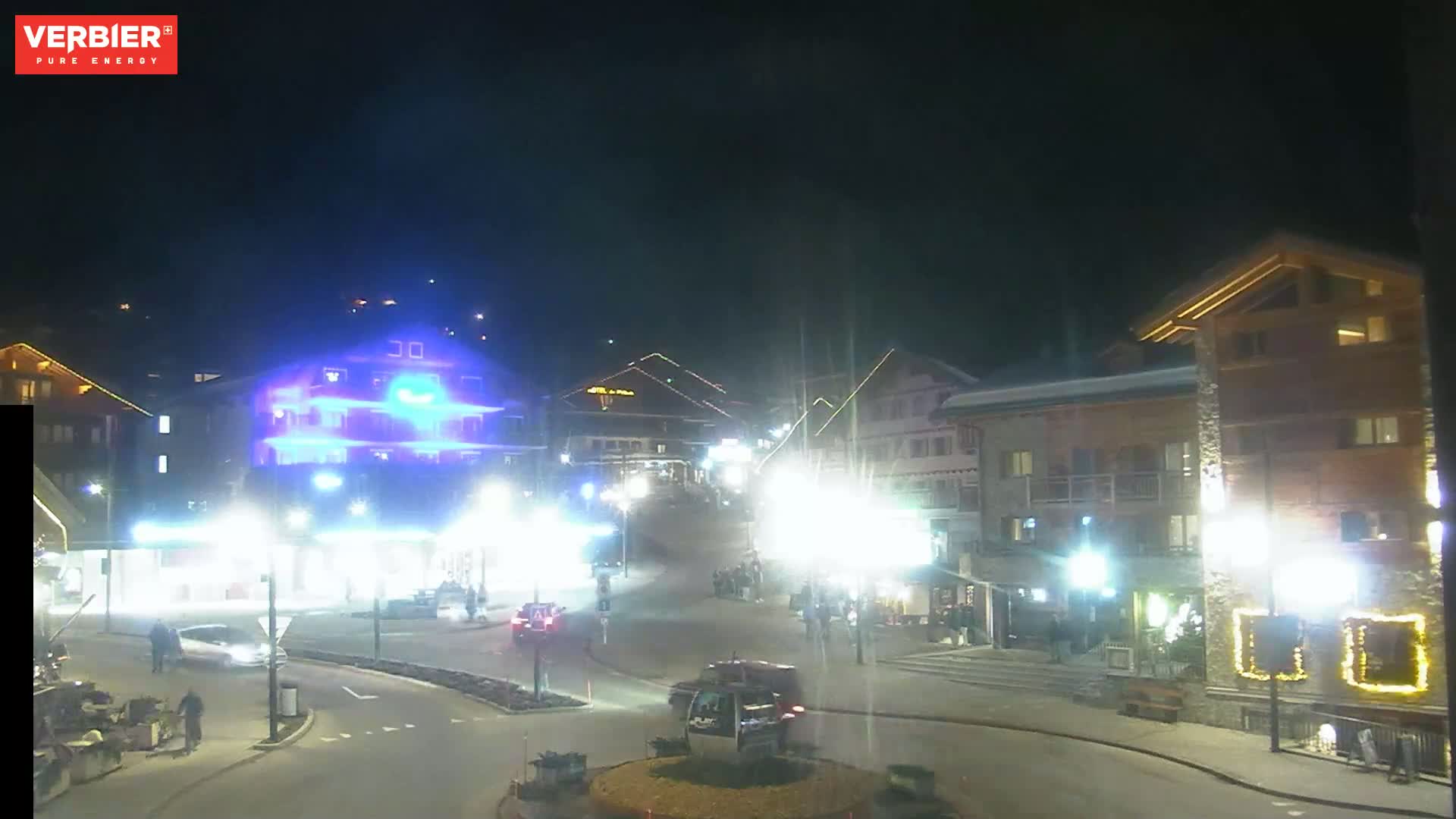A nighttime view of a town square with illuminated buildings and cars, under a dark sky.