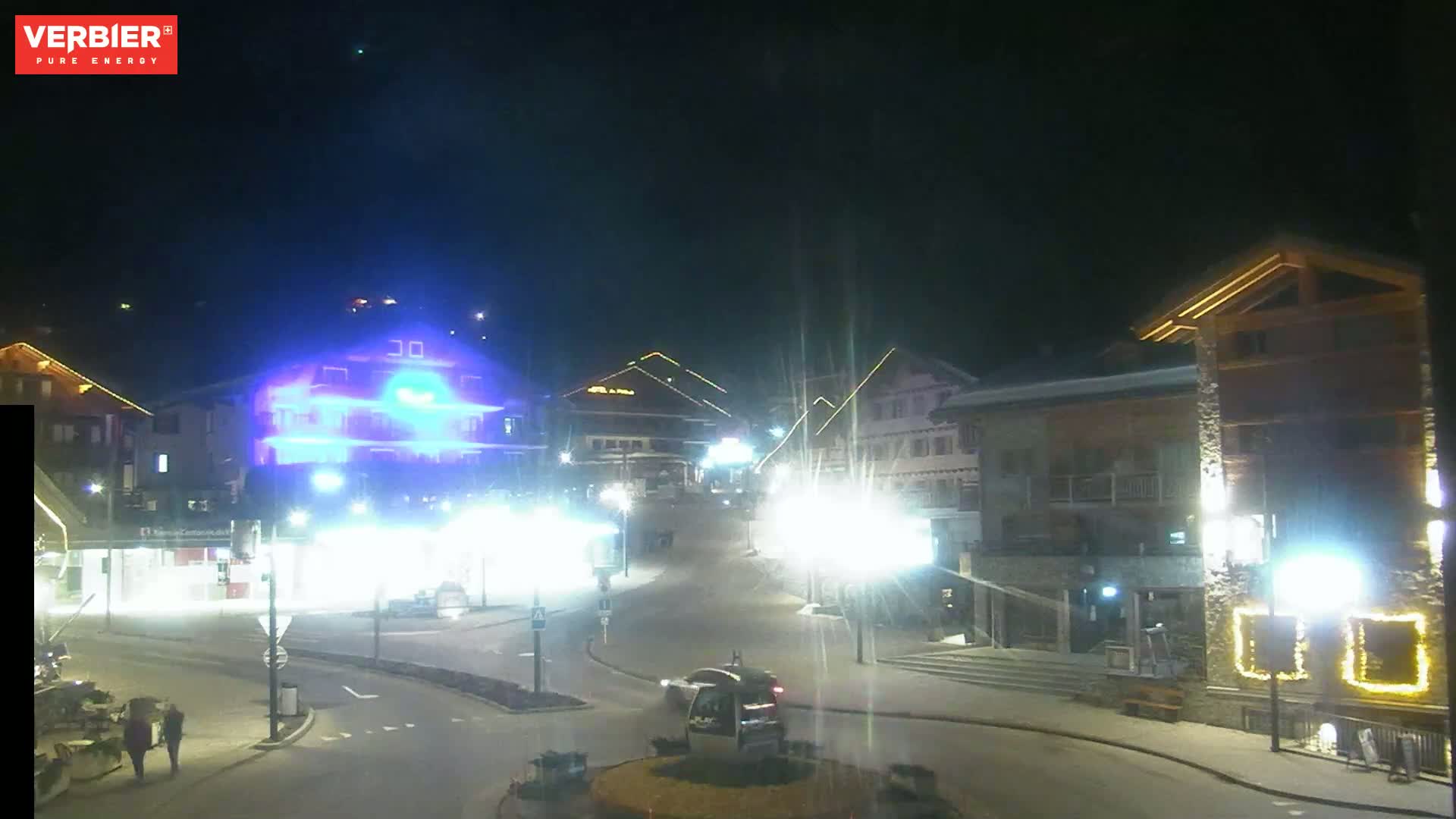 A nighttime view of a snow-covered town square with brightly lit buildings and a few cars, under a dark, clear sky.