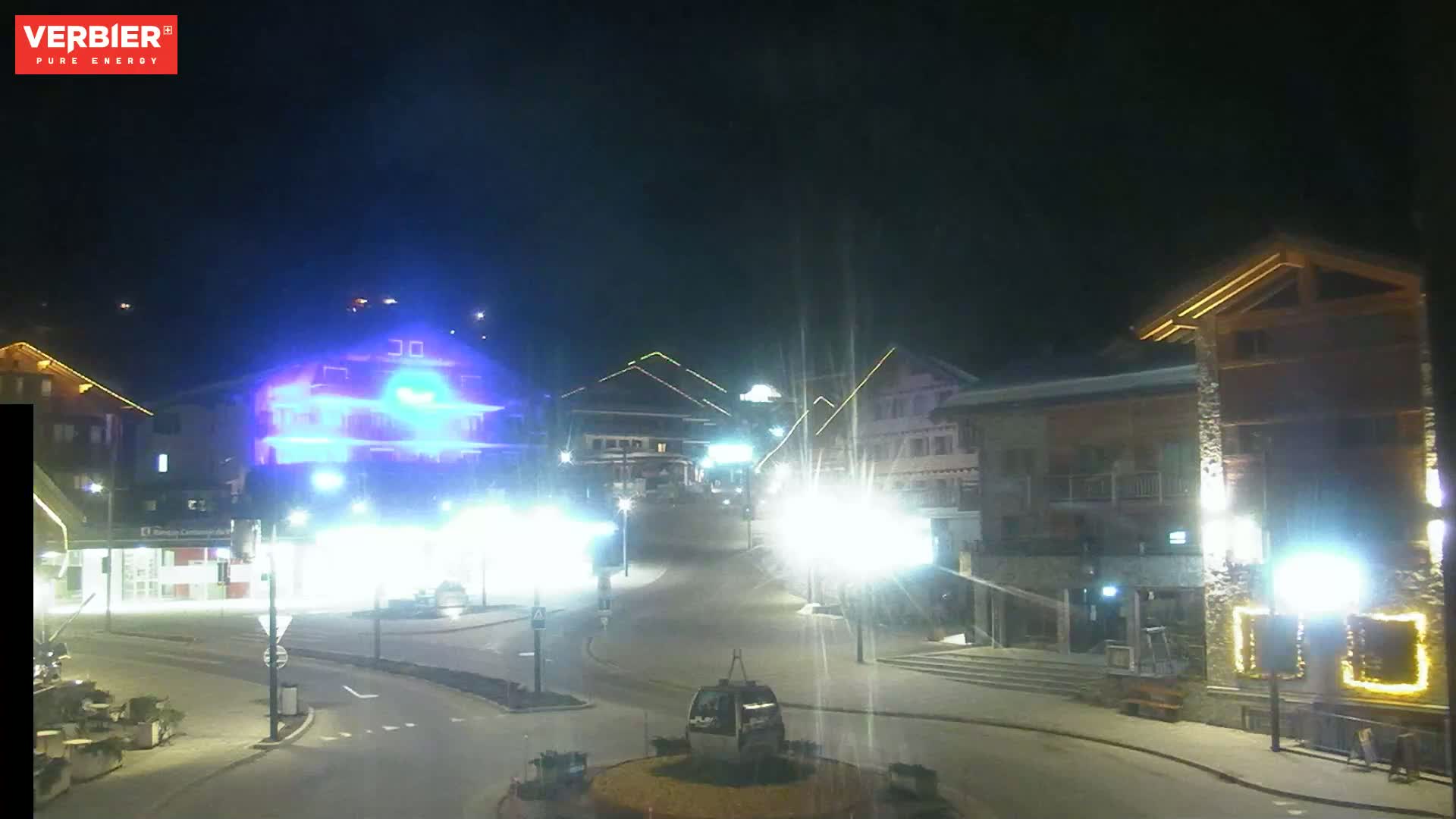 A nighttime view of a town square with several buildings illuminated, a gondola lift in the center, and bright streetlights.