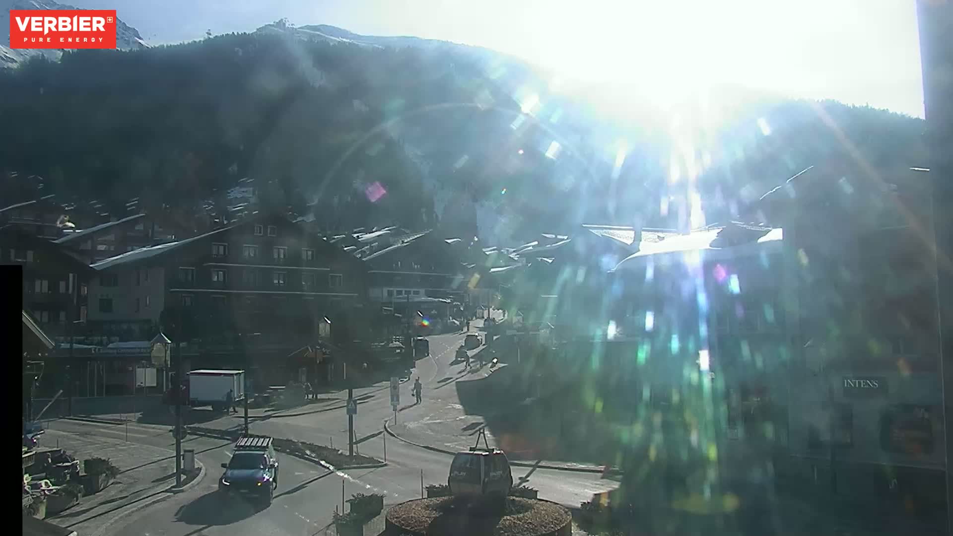A sunny, bright day in a mountain village shows a street scene with buildings, vehicles, and a gondola lift, with a mountain backdrop.