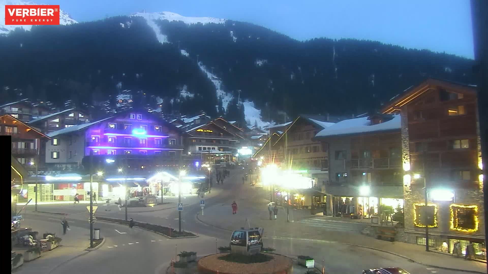 A nighttime view of a snow-dusted mountain village square, with illuminated buildings and a few pedestrians, under a twilight sky.