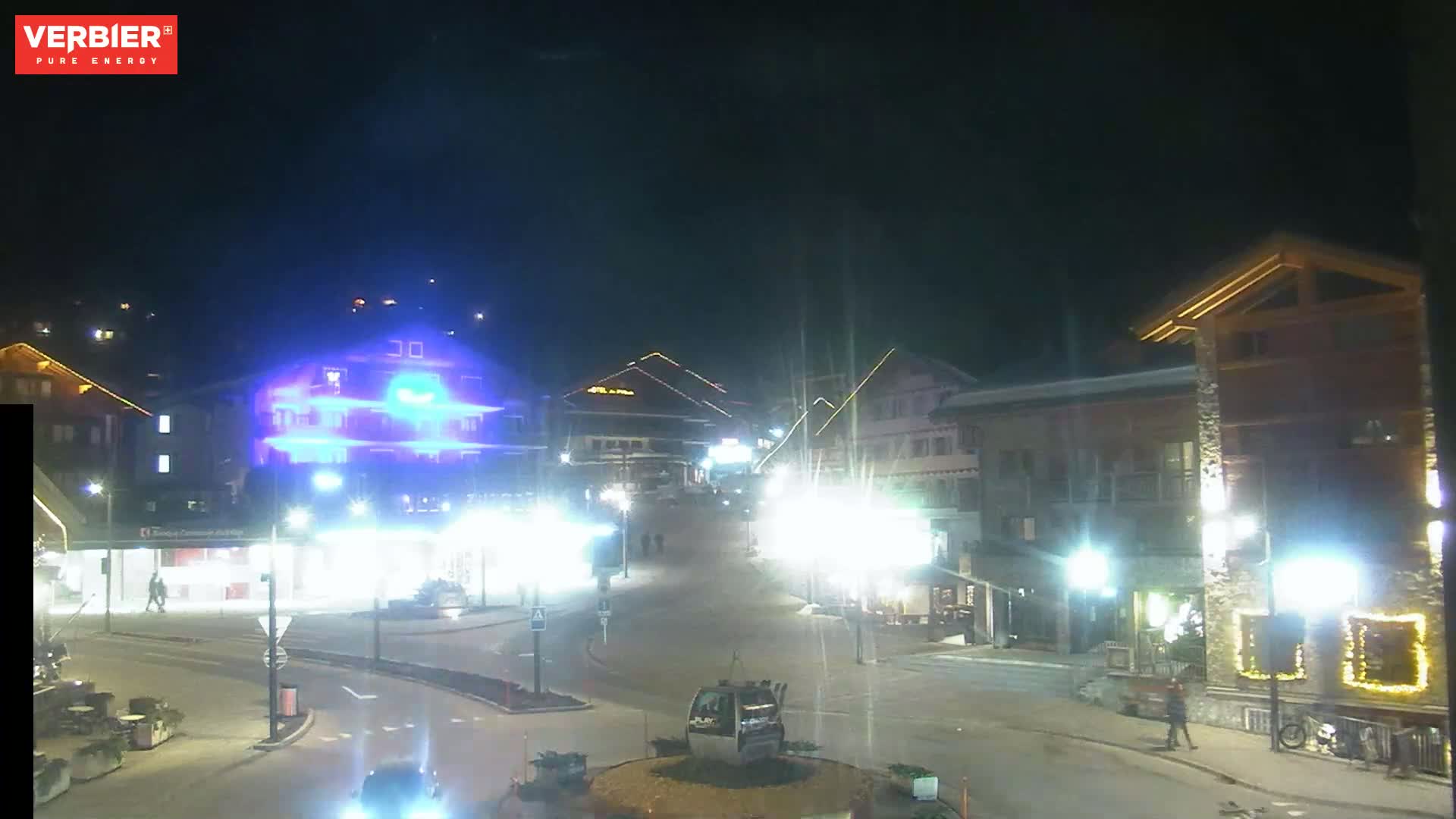 A nighttime view of a town square with brightly lit buildings, streetlights, and a few pedestrians, under a dark sky.