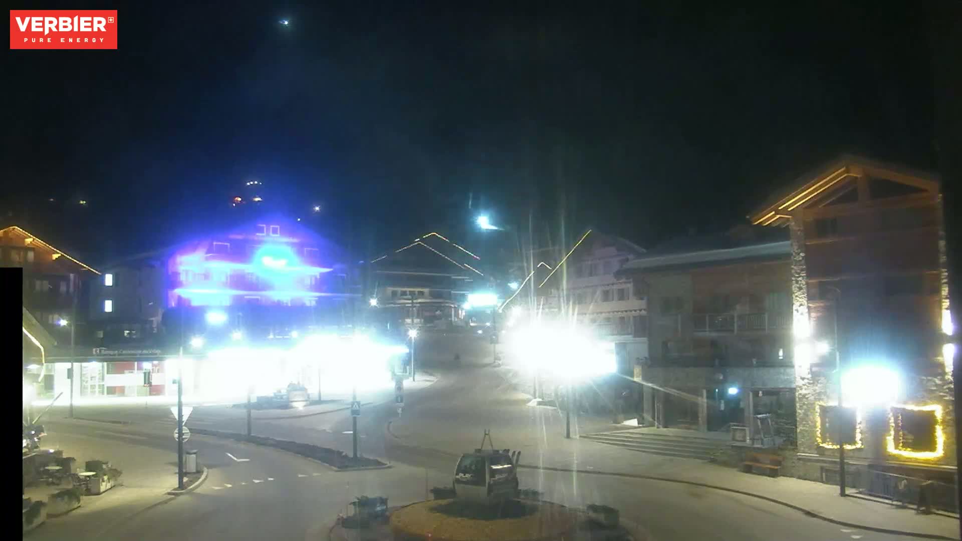 A brightly lit town square at night, with several buildings illuminated, a cable car visible in the foreground, and a clear night sky.
