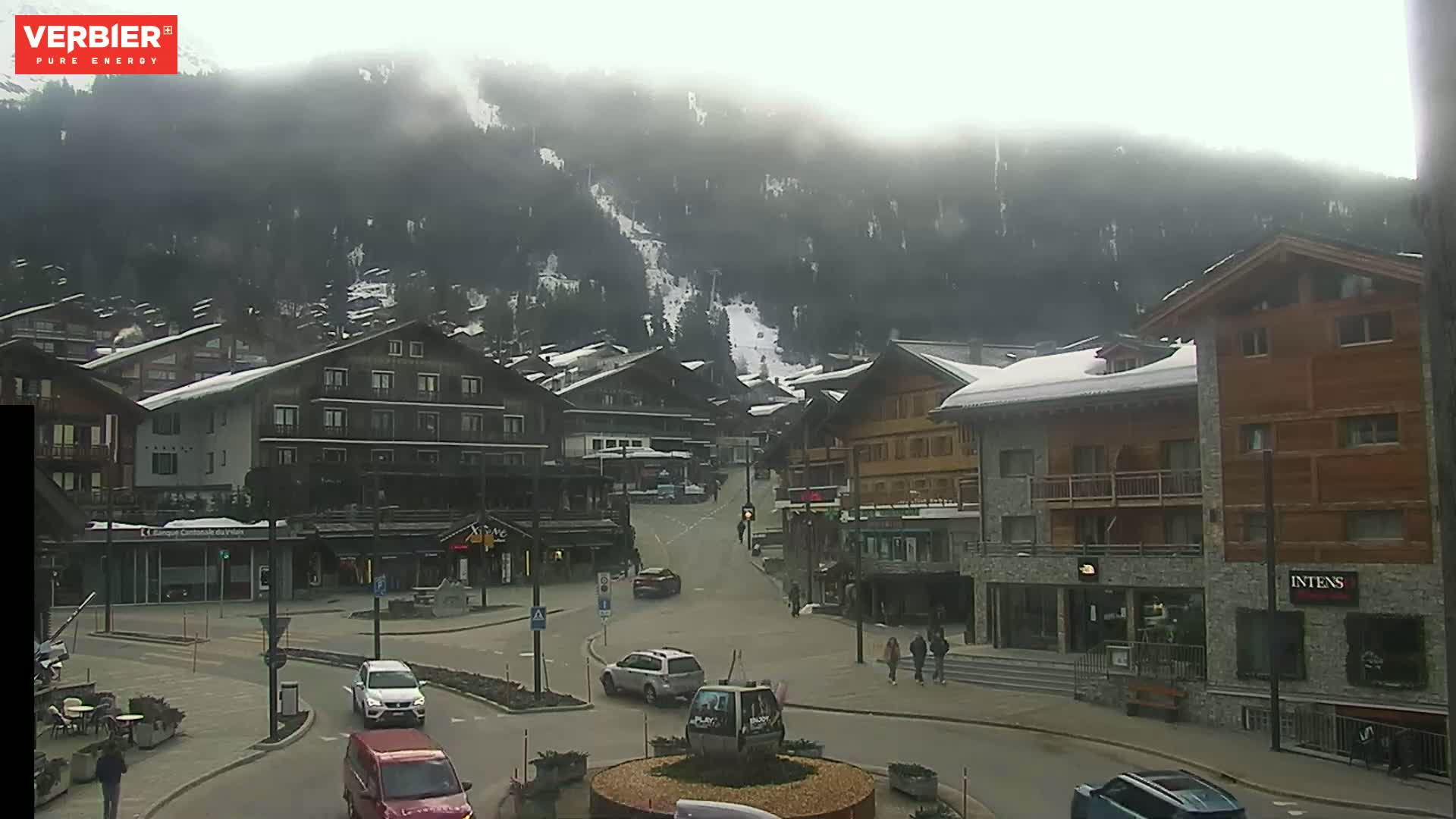 A foggy day reveals a snow-dusted alpine village nestled at the base of a mountain with a ski slope visible, and several cars are parked or driving on the street.